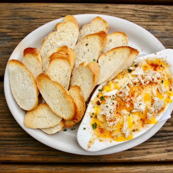 A white plate with toasted baguette slices and a dish of melted cheese dip with crab meat.