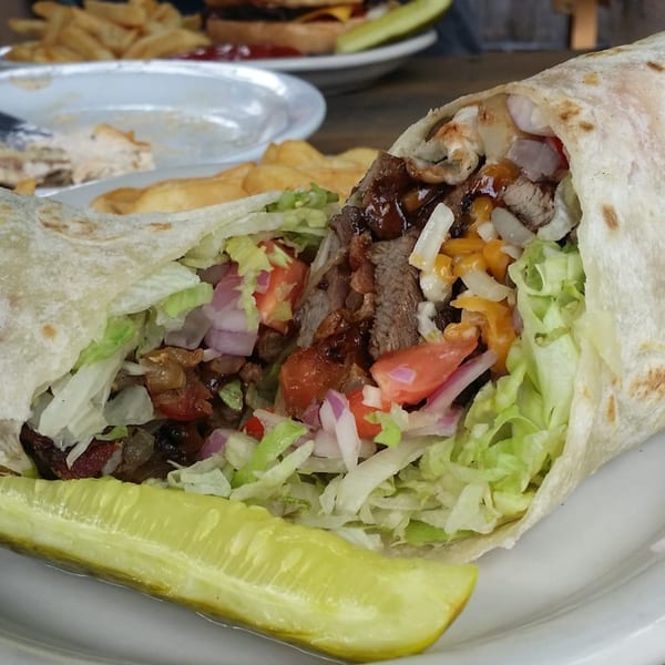 A Stan & Joe's Saloon steak wrap, filled with meat, lettuce, tomatoes, onions, and cheese, with a pickle spear.