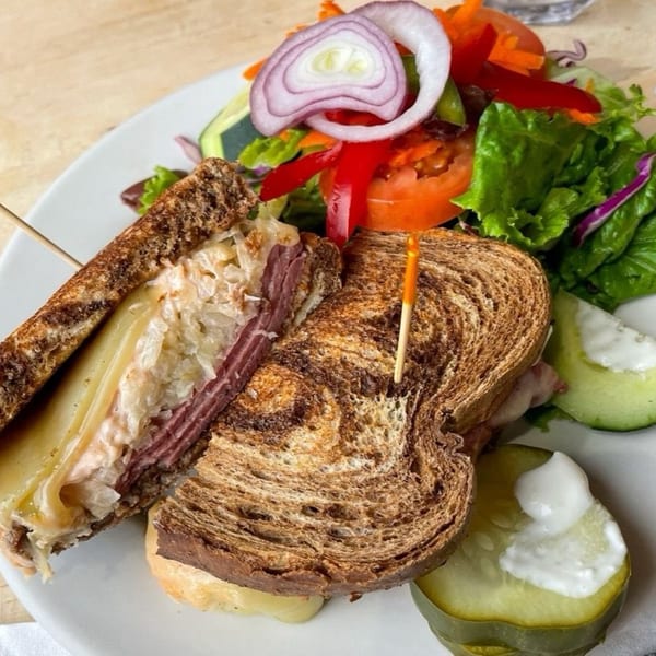 A Stan & Joe's Saloon Reuben sandwich with corned beef, Swiss cheese, sauerkraut, and Russian dressing, served with a side salad and pickles.