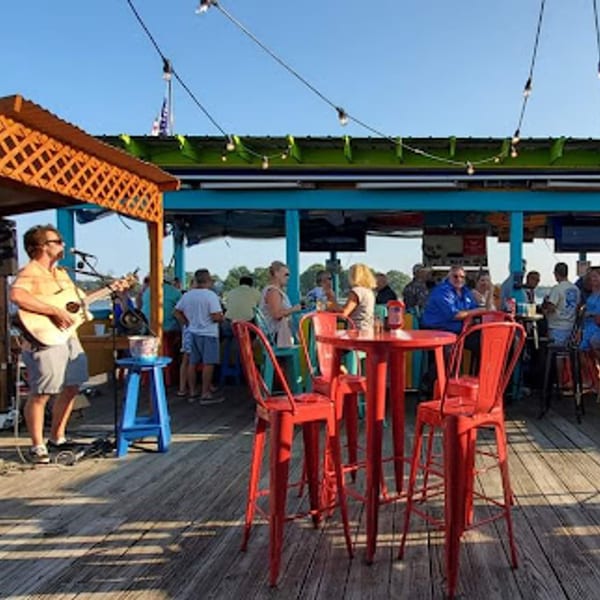 A musician plays guitar on a wooden deck with red bar stools and people socializing at Stan & Joe's Saloon.