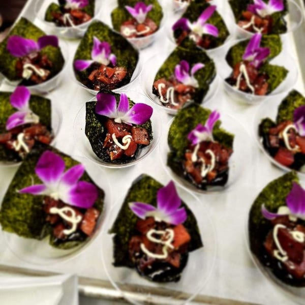 Numerous small appetizers with tuna, seaweed, white sauce, and purple orchid garnishes on a tray.